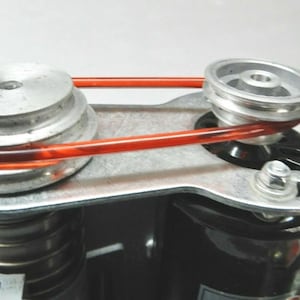 Może przedstawiać: Zbliżenie na srebrny metalowy system rolek z czerwonym gumowym paskiem. System rolek jest przymocowany do czarnego silnika.