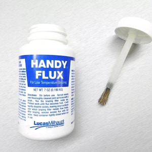 Puede incluir: Una botella blanca con una etiqueta azul que dice "HANDY FLUX" para soldadura de baja temperatura. La botella contiene 7 onzas (0,190 kg) de fundente y tiene una tapa blanca con un aplicador de cepillo.