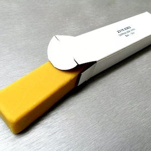 Op de afbeelding: Een witte kartonnen doos met de tekst "2015-2022 Carnauba Wax Bar 2oz" gedrukt op de bovenkant. De doos is open en een gele wasbalk is gedeeltelijk zichtbaar in de doos.