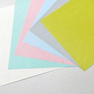 Peut inclure: Une pile de cinq feuilles de papier de différentes couleurs, dont blanc, bleu clair, rose, vert clair et jaune. Les feuilles sont disposées en éventail, la feuille blanche en bas et la feuille jaune en haut.