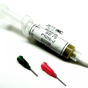 May include: A clear syringe filled with a light brown substance, labeled "SS 75 P101E-2 B7708" from JETS INC. Two colored applicator tips, one green and one red, are positioned below the syringe.
