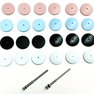 May include: A collection of dental polishing wheels in various colors, including pink, blue, black, and white. The wheels are arranged in rows and columns, with two metal mandrels at the bottom. The wheels have a small hole in the center.