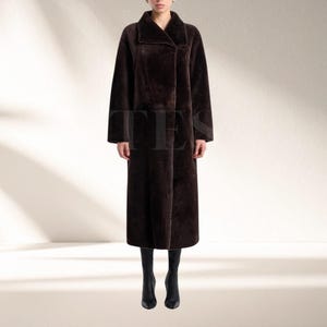 May include: A full-length, dark brown coat with a wide collar and button closure. The coat is made of a soft, textured material. It is worn with black boots. The coat's length extends to the ankles. The style is elegant and suitable for colder weather.