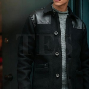 Könnte beinhalten: Ein schwarzer Wollmantel mit Lederbesätzen an Schultern und Kragen. Der Mantel hat eine Knopfleiste, zwei Vordertaschen und ein klassisches Design. Das Model trägt einen grauen Pullover darunter.