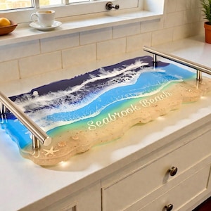 May include: A resin serving tray with a beach design. The tray has a blue and white wave pattern, a sandy beach area, and the text "Seabrook Beach." It has silver handles. The tray is approximately 60 cm long.