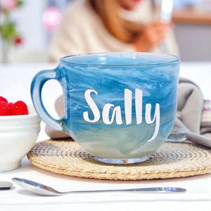 May include: A blue and white marbled glass mug with the name "Sally" in white script. The mug sits on a woven coaster, with a small white bowl of red berries and a spoon in the foreground. The mug has a large handle.