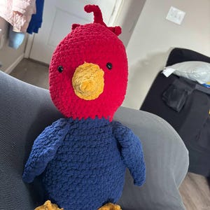 Op de afbeelding: Een handgemaakte gehaakte vogelknuffel met een rode kop, gele snavel en blauw lichaam. De vogel heeft kleine zwarte ogen en gele poten. Dit zachte speelgoed is een uniek, handgemaakt item.