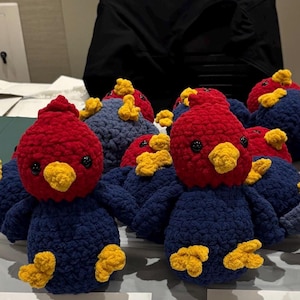 May include: A collection of small, handmade crochet birds featuring dark blue bodies, bright red heads, yellow beaks, and yellow feet. Each bird has two small black plastic eyes. The birds are arranged in a group, showcasing their soft, textured yarn construction.