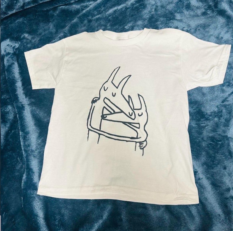 Puede incluir: Camiseta blanca con un dibujo lineal negro de dos figuras abstractas abraz&aacute;ndose. El gr&aacute;fico est&aacute; centrado en la parte delantera de la camiseta. La camiseta tiene mangas cortas y cuello redondo.