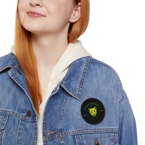 May include: A light blue denim jacket with a round black patch featuring a green and yellow panther design and the word "CENTENNIAL". The jacket is worn over a cream-colored hoodie.