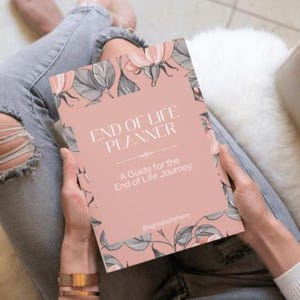 Könnte beinhalten: Ein rosa "End of Life Planner" Buch mit floralen Illustrationen wird von einer Person gehalten, die zerrissene Jeans trägt. Das Cover des Buches zeigt den Titel und den Untertitel "A Guide for the End of Life Journey". Das Buch wird auf einer weißen, flauschigen Oberfläche gehalten.