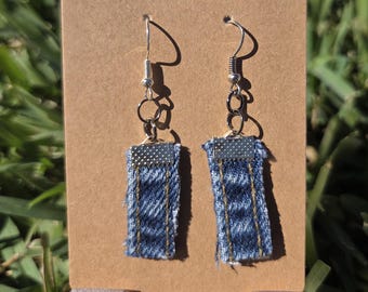 Op de afbeelding: Een paar handgemaakte denim oorbellen. Elke oorbel heeft een rechthoekig stuk blauwe denimstof met gerafelde randen, geaccentueerd met zilverkleurige metalen fournituren. De oorbellen hangen aan zilverkleurige oorhaken.