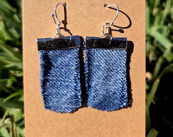 Op de afbeelding: Een paar denim oorbellen met zilverkleurige metalen accenten. De oorbellen zijn rechthoekig van vorm en hebben een donkerblauwe denim stof. De bovenkant van elke oorbel heeft een zilverkleurige metalen staaf en een zilverkleurige haak.