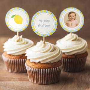 Könnte beinhalten: Drei Cupcakes mit weißem Zuckerguss und dekorativen Toppern. Einer zeigt eine gelbe Zitrone, ein anderer liest "my zesty first year", und der dritte hat ein Babyfoto. Die Cupcakes stehen auf einer Holzoberfläche.