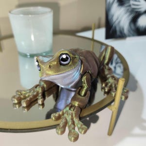 May include: A 3D printed frog figurine with a green and brown body and large, round eyes. The frog is sitting on a gold-colored, round, mirrored tray with a gold stand.
