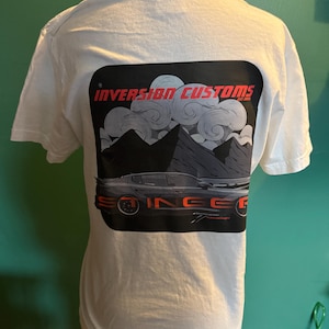 Puede incluir: Camiseta blanca con un diseño gráfico en la espalda. El diseño incluye el texto "INVERSION CUSTOMS" en rojo, un coche, montañas y nubes. La palabra "SO INGE" también es visible en rojo.