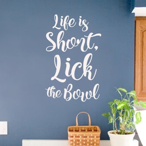 May include: White vinyl wall decal with the text "Life is Short, Lick the Bowl" on a blue wall.