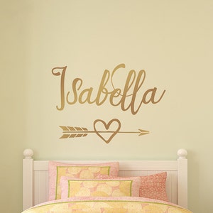 May include: Gold vinyl wall decal with the name "Isabella" in a cursive font, an arrow with a heart in the middle, and a white headboard behind it.