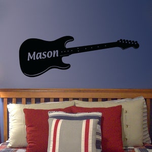 Puede incluir: Pegatina de pared de guitarra eléctrica negra con el nombre "Mason" en el cuerpo de la guitarra. La pegatina está sobre una cabecera de madera con una cama con almohadas y una colcha.