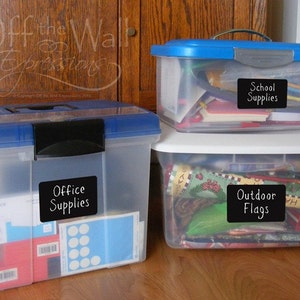 May include: Three clear plastic storage bins with black lids. The bins are labeled with black chalkboard labels. The top bin is labeled "School Supplies", the middle bin is labeled "Office Supplies", and the bottom bin is labeled "Outdoor Flags".