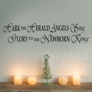 Puede incluir: Una pared blanca con letras negras en cursiva que dicen "Hark the Herald Angels Sing Glory to the Newborn King". Hay un pequeño árbol de Navidad de alambre verde con bayas rojas y una estrella en la parte superior. Hay cuatro velas blancas sobre una superficie de madera.