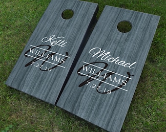 May include: Two wooden cornhole boards with a gray woodgrain finish. The boards are personalized with the names "Kelli" and "Michael" along with the last name "Williams" and the date "7.27.19".