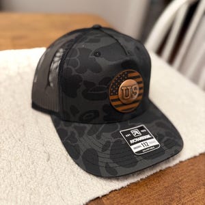 May include: Black camouflage trucker hat with a curved brim and mesh back. Features a copper-colored patch with "US" and a flag design. The hat has a "Richardson" tag and is adjustable.