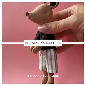 Pode incluir: Um brinquedo de pelúcia com cabeça bege, orelhas castanho-escuras e nariz castanho-escuro. O brinquedo veste uma camisa preta de manga comprida e calções às riscas brancas e verdes com um pequeno laço branco. Texto: PDF SEWING PATTERN THE ESSENTIAL T-SHIRT.
