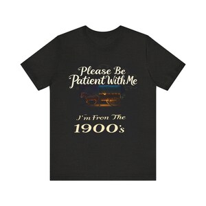 May include: Dark heather gray t-shirt featuring a vintage-style design. The text reads, 'Please Be Patient With Me I'm From The 1900's.'  An image of a horse-drawn wagon is displayed above the text. The design is in cream-colored lettering against a dark background.