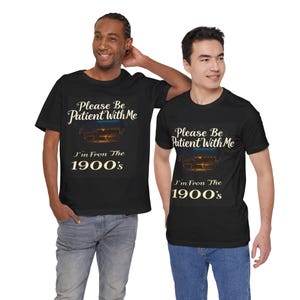 May include: Two black t-shirts with a vintage-style design. The shirts feature the text 'Please Be Patient With Me, I'm From The 1900's' and an image of a horse-drawn carriage. The text is in a cream color against the black background. The shirts are shown on two models.