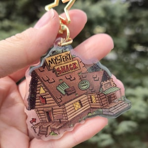 May include: A clear acrylic keychain featuring a cartoon illustration of the Mystery Shack, with the text "MYSTERY SHACK" on a sign. The keychain has a gold star-shaped clasp.