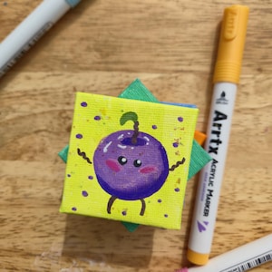 May include: A small, square canvas painting featuring a cartoon blueberry character with a smiling face, arms, and legs. The background is yellow with purple dots. The painting is on a green square, with a wooden surface and markers in the background.