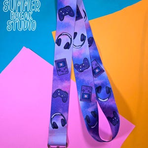 May include: A purple lanyard with a galaxy design featuring video game controllers, headphones, and a handheld console. The lanyard has a silver clasp and the text "SUMMER BREAK STUDIO" in white.