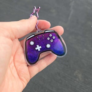 May include: A purple and blue glittery video game controller keychain with white buttons and a plus sign. The keychain has a clear border and a star-shaped clasp.