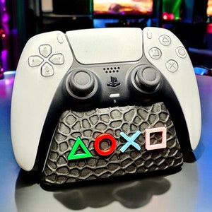 Può includere: Un controller PlayStation 5 DualSense bianco con una base testurizzata nera e grigia. La base ha un triangolo verde, un cerchio rosso, una X blu e un quadrato rosa.