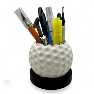 Peut inclure: Un organiseur de bureau en forme de balle de golf blanche avec une base noire, contenant divers stylos, marqueurs et un cutter. L'organisateur a une surface à fossettes, ressemblant à une vraie balle de golf. Les articles à l'intérieur sont de couleurs différentes.