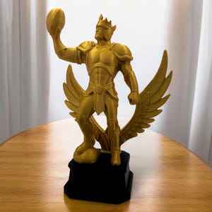 Puede incluir: Estatua dorada de una figura sosteniendo un balón de fútbol americano, con corona y alas. La estatua está montada sobre una base negra. La figura lleva armadura y está en una pose heroica. Las alas están extendidas.