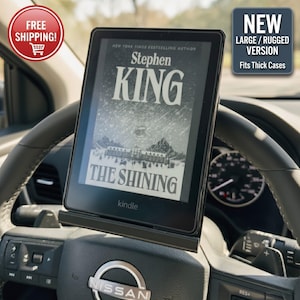 Puede incluir: Lector de libros electrónicos Kindle negro que muestra el libro "The Shining" de Stephen King, montado en el volante de un coche. El lector está en un soporte negro. Incluye el texto "NEW LARGE/RUGGED VERSION Fits Thick Cases".