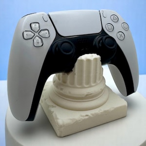 Pode incluir: Um comando de jogo PlayStation 5 DualSense branco é exibido em um suporte branco impresso em 3D com a forma de uma coluna grega. O suporte tem uma base quadrada.