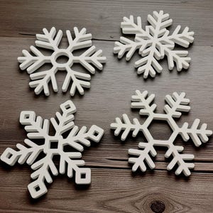 May include: Four white snowflake-shaped decorations with intricate designs. Each snowflake has a unique pattern of arms and details, perfect for winter or holiday decor. The snowflakes are displayed on a dark wooden surface.