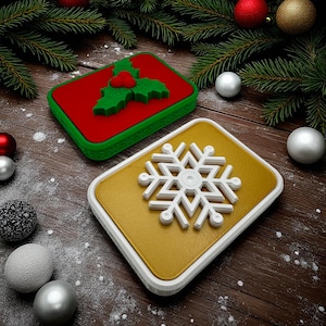 May include: Two Christmas-themed decorative boxes. One is red with a green holly design, the other is gold with a white snowflake. Both have white and green borders. The boxes are on a wooden surface with Christmas ornaments and pine branches.