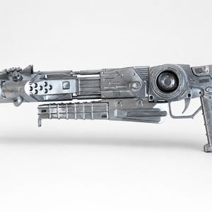 May include: A silver-colored sci-fi blaster prop featuring a long barrel with circular vents, a textured grip, a trigger, and a complex mechanical design with various panels, ridges, and a circular side component, presented against a plain background.