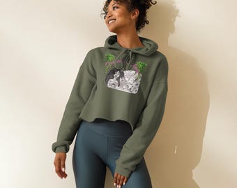 Op de afbeelding: Een olijfgroene cropped hoodie met een trekkoord en lange mouwen. De hoodie heeft een grafische print van twee vrouwen en bloemelementen. Het model draagt ook een donkerblauwe sportbroek.
