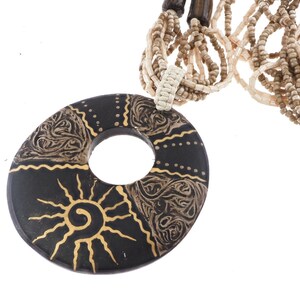 May include: A multi-strand necklace featuring a large, circular black pendant with gold-painted sun and swirl motifs. The necklace is composed of numerous thin strands of small, light beige and tan glass beads, connected by a woven cream-colored cord.