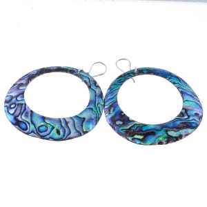 May include: A pair of large, circular hoop earrings crafted from iridescent abalone shell. The surface displays a swirling, natural pattern of deep blues, vibrant teals, and hints of purple. Each earring features a simple silver-toned wire hook.