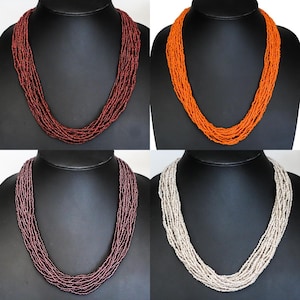 Puede incluir: Cuatro collares de cuentas en diferentes colores: burdeos, naranja, malva y blanco roto. Cada collar está hecho de múltiples hebras de pequeñas cuentas, exhibidas en un busto de maniquí negro. Los collares son una pieza llamativa.