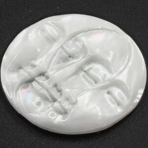 May include: A white, round, ceramic moon with a face sculpted into the surface. The face has a detailed expression with eyes, nose, and mouth.