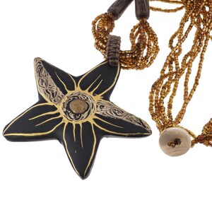 May include: A multi-strand necklace featuring a large, black, five-pointed star pendant with gold-painted floral details and a central metallic accent. The necklace consists of numerous strands of small, metallic bronze beads and a single white button.