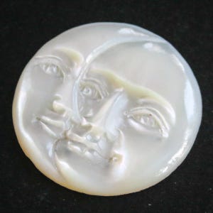 Op de afbeelding: Een witte parelmoer cameo-gravure met twee gezichten, één aan elke kant van de maan, met gedetailleerde kenmerken.
