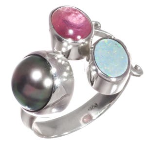 May include: A silver ring featuring three gemstones: a large, dark gray pearl, a pink gemstone, and a light blue opal. The gemstones are set in individual silver bezels, creating a unique and eye-catching design. The ring has a modern, artistic style.